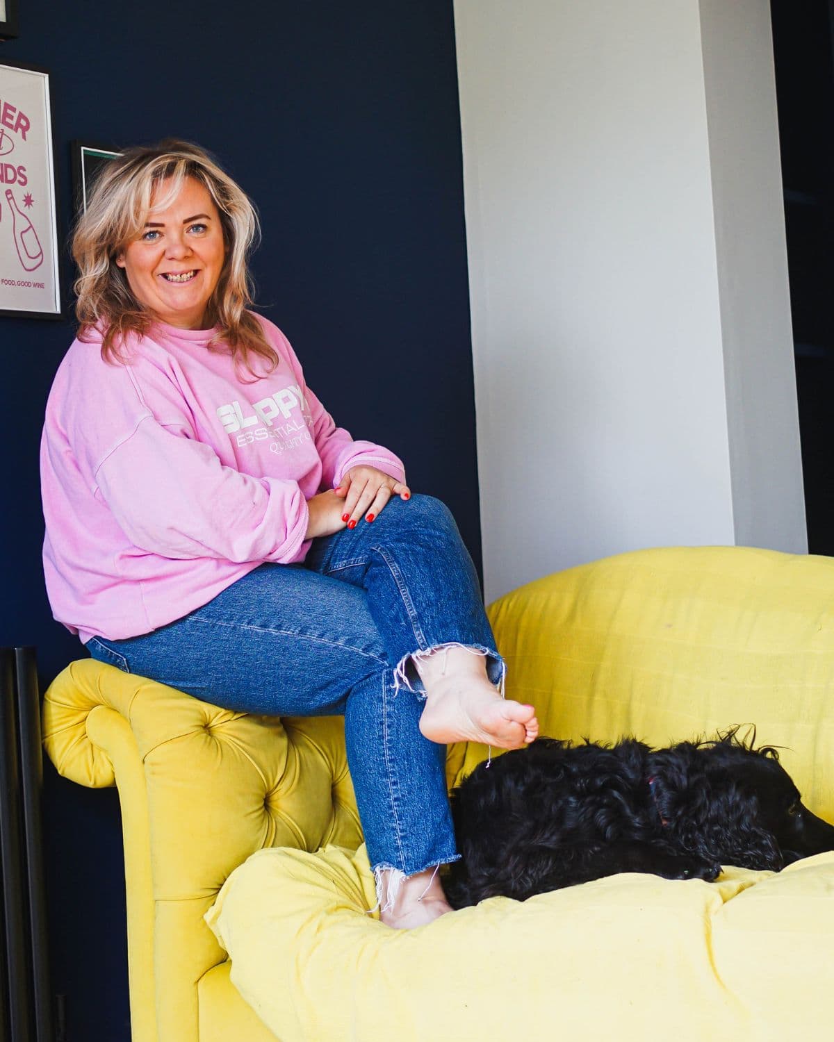 Zoë Dew sitting on the arm of a yellow sofa in front of a navy wall, smiling, with her dog asleep beside her