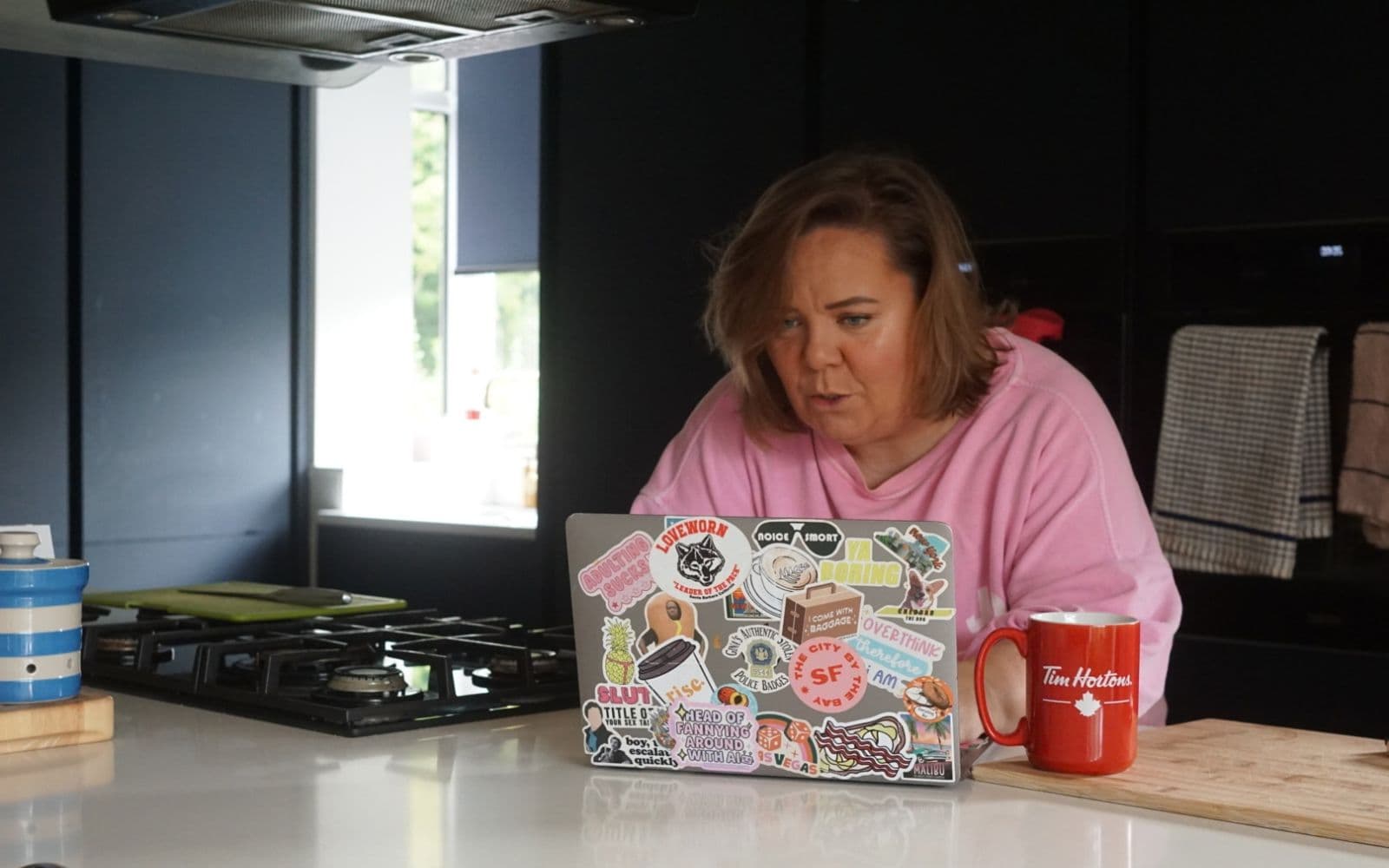 Zoë Dew at her kitchen counter, concentrating hard on her sticker-covered laptop, with a red mug beside her