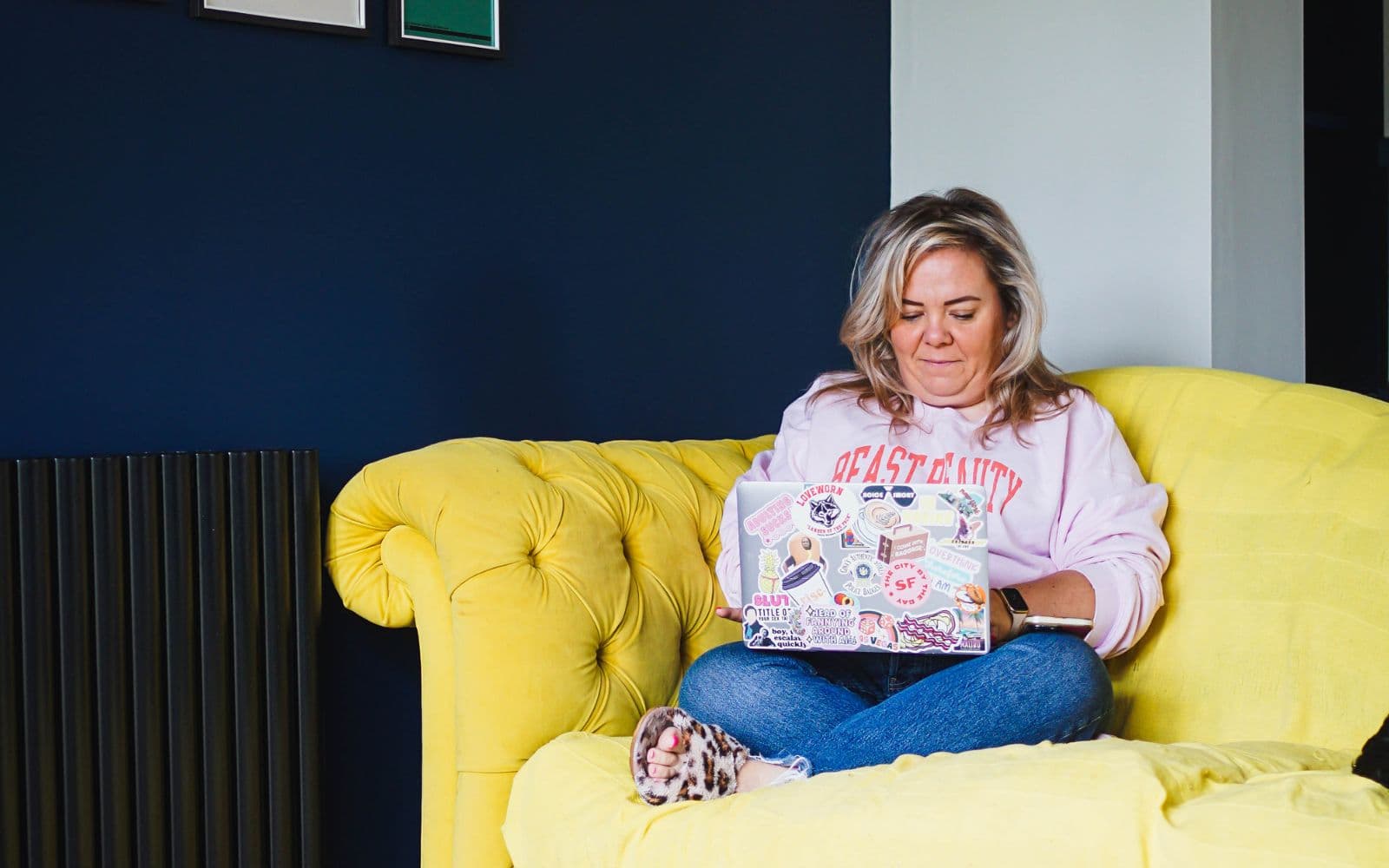 Blonde woman (Zoë Dew) sat in a living room on yellow sofa working on her laptop not looking at the camera