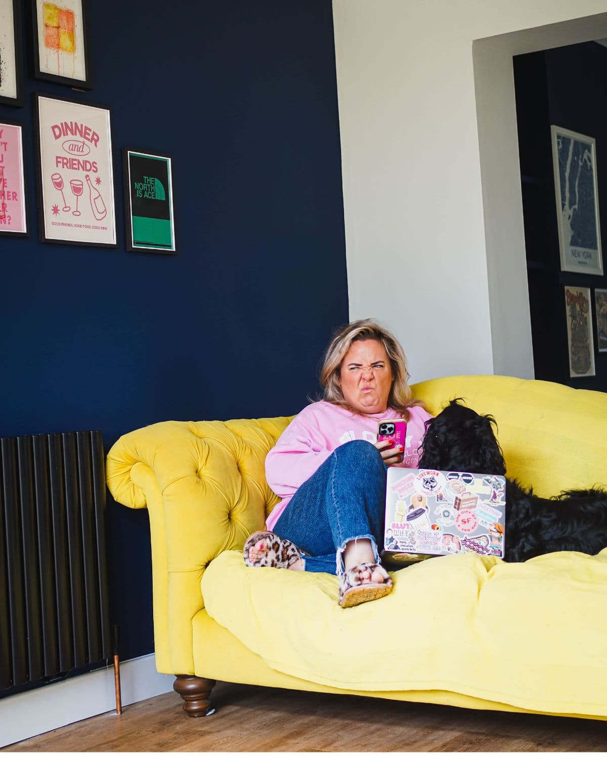Zoë Dew on a yellow sofa with her phone, laptop and dog, pulling an exasperated face