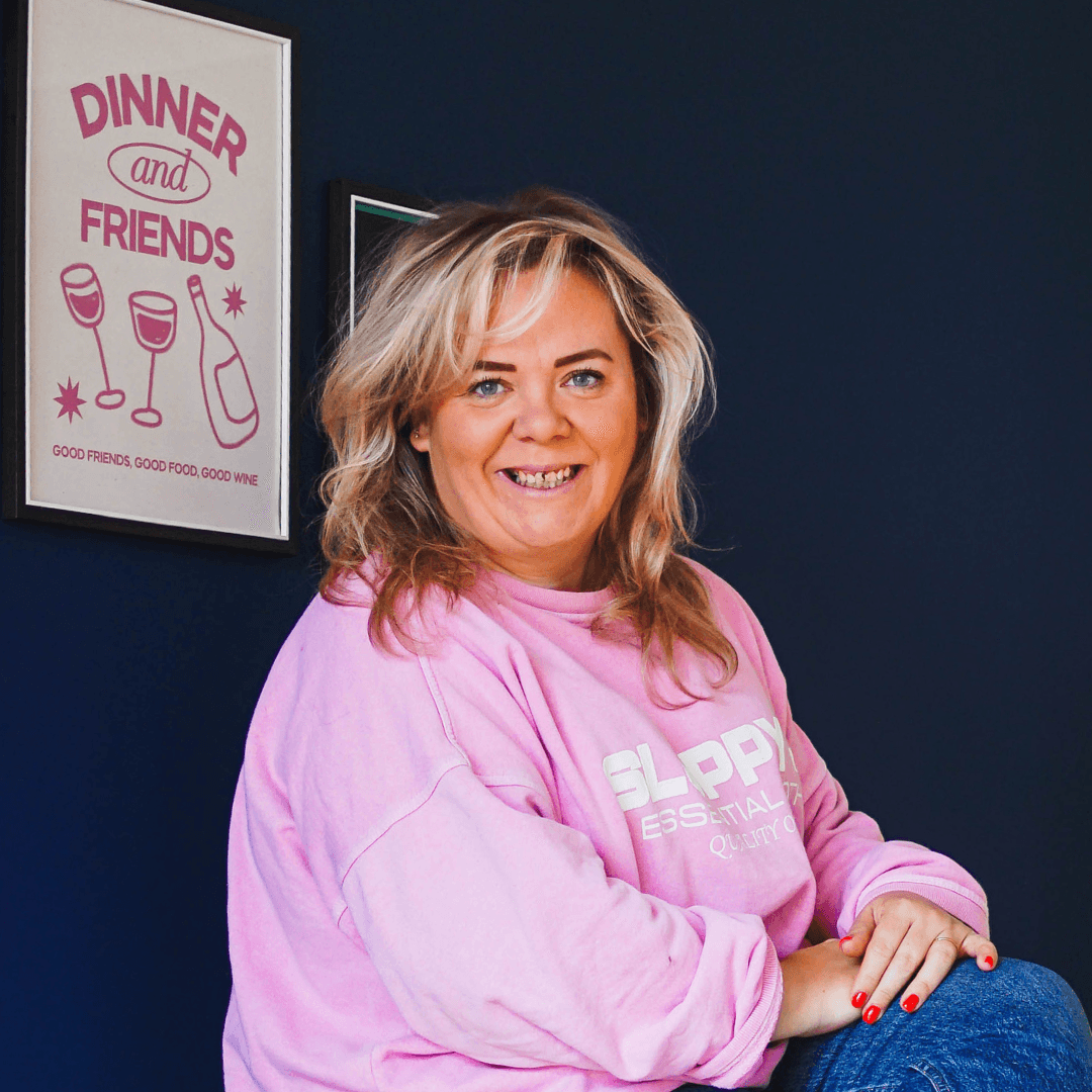 Zoë Dew smiling, in a pink sweatshirt in front of a navy wall with a framed print