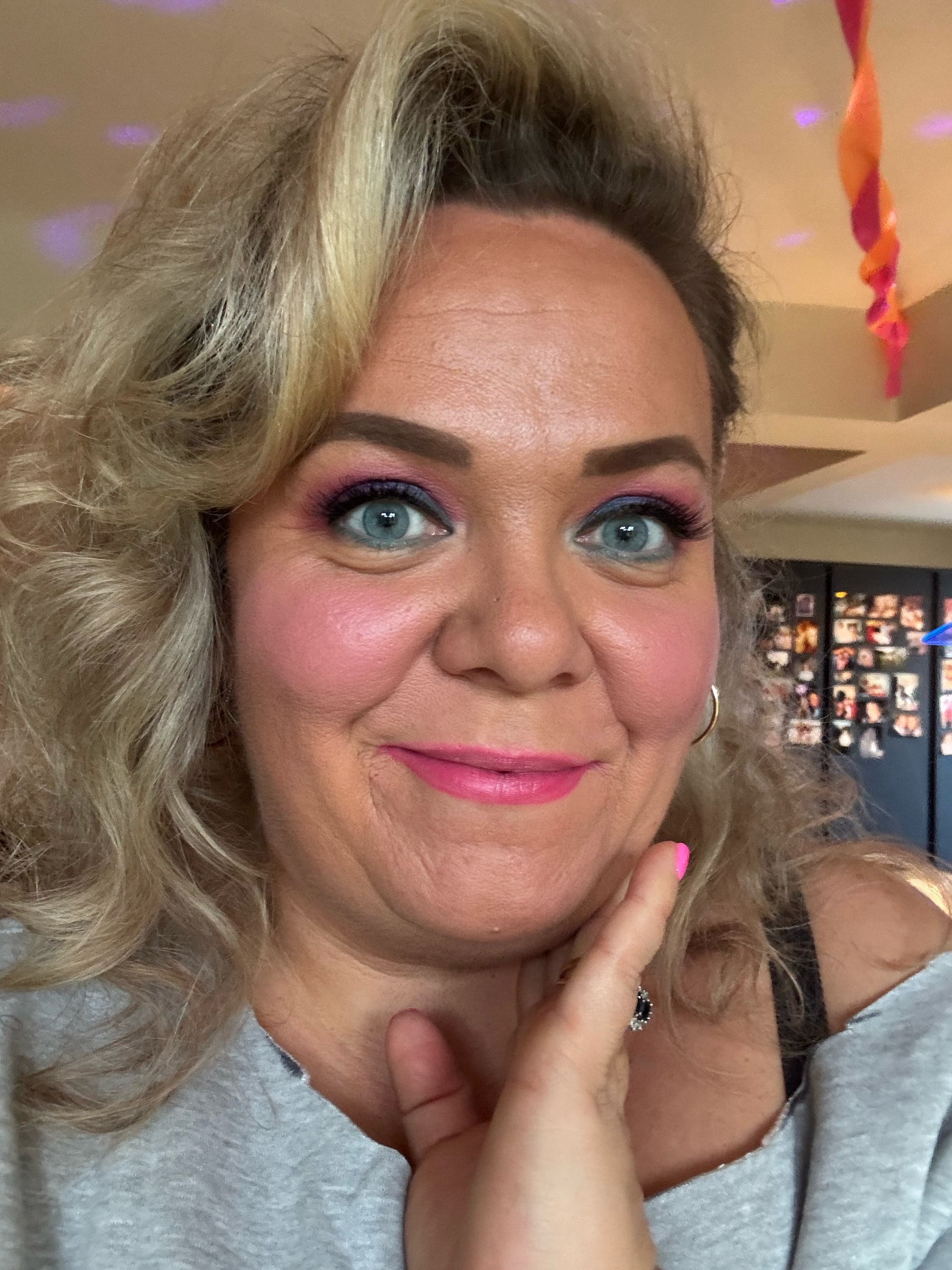 A white woman with blonde hair portrait her hand is to her face and she has bright eyeshadow and lipstick